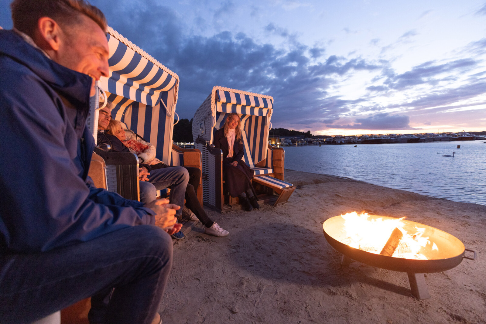 Menschen sitzen in Strandkörben bei Sonnenuntergang mit Feuerschale.