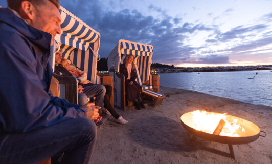 Menschen sitzen in Strandkörben bei Sonnenuntergang mit Feuerschale.