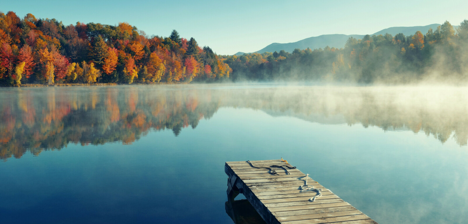Ein einsamer Steg ragt in einen nebeligen See, am anderen Ufer erkennt man einen Herbstwald.
