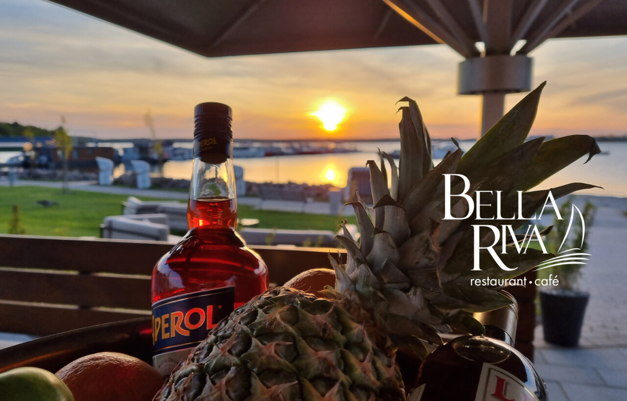 Aperol und Ananas vor einem Sonnenuntergang am Yachthafen im MareMüritz mit dem Logo des Bella Riva Restaurants.