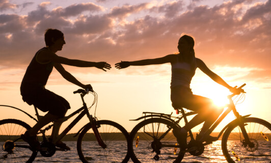 Ein Pärchen fährt am Seeufer vor einem Sonnenuntergang gemeinsam Fahrrad.